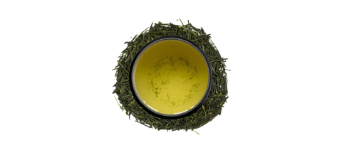 A top-down view of a black ceramic bowl filled with pale yellow-green tea, surrounded by loose green tea leaves on a plain white background.