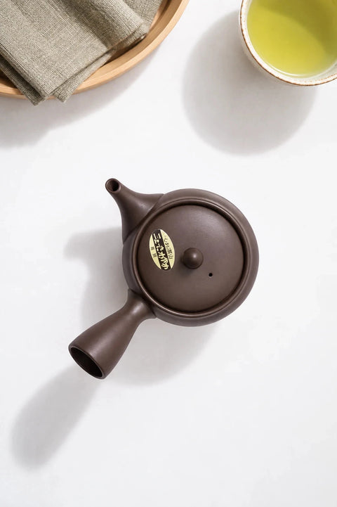 A dark brown ceramic teapot with a gold emblem on the lid, styled on a light beige stone surface with natural leaf shadows and tea accessories nearby.