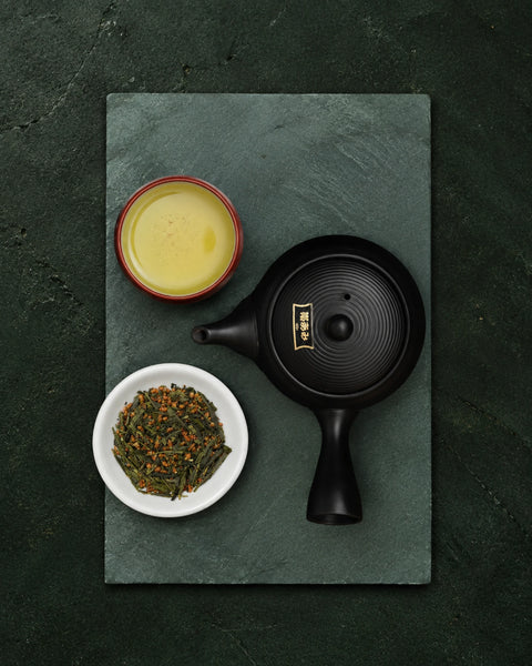 A black ceramic teapot with gold characters on the lid, arranged on a dark green slate tray with a cup of yellow tea and a small bowl of genmaicha loose leaf tea.