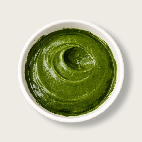 A top-down view of a thick, vibrant green puree, likely avocado or pumpkin puree, swirled in a white ceramic bowl against a light gray background.
