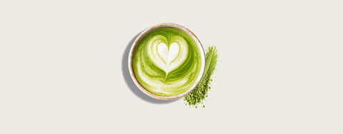 A top-down view of a matcha latte in a small ceramic cup, featuring a heart-shaped latte art design in green and white. A small pile of loose green matcha powder sits beside the cup on a light beige background.