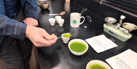 Nio Teas team member is holding a small spoon with green matcha powder over a small bowl, with other matcha-related items including a white mug with a green logo, a balance scale, and printed papers on a black tabletop.