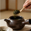 A close-up of a dark brown ceramic teapot with a hand holding a wooden spoon filled with loose green tea above it, set on a light tabletop in a softly blurred indoor setting.