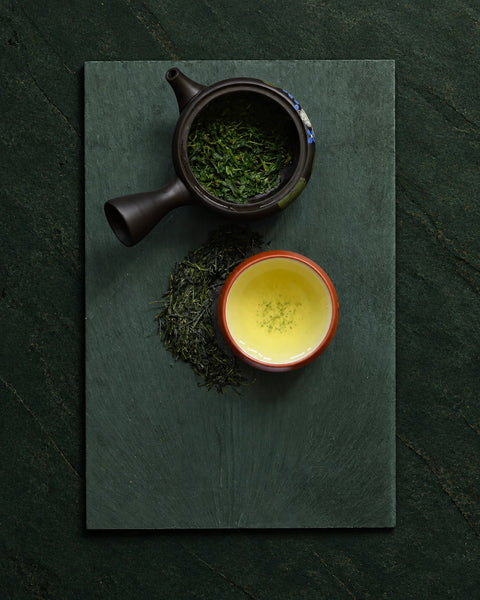 A dark ceramic teapot filled with gyokuro loose green tea leaves and a small cup of brewed yellow-green tea, arranged on a dark green rectangular board against a textured dark green background.