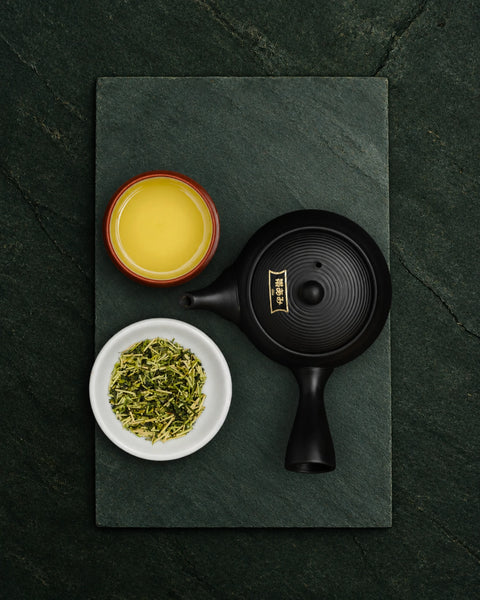 A top-down flat lay of a black ceramic teapot with a gold emblem, a small bowl of kukicha tea, and a small dish of loose green tea leaves, arranged on a dark green slate board over a dark textured background.