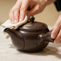A close-up of a dark brown ceramic teapot with a person's hands cleaning the lid with a white cloth, set against a warm beige background.