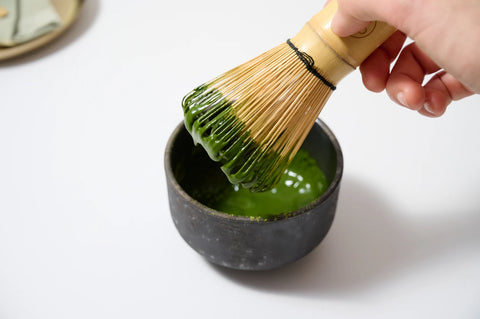 A hand holds a bamboo whisk above a small dark bowl filled with bright green matcha tea on a clean white surface.