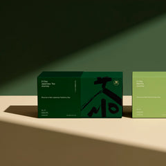 Two rectangular product boxes on a beige surface against a dark green background, lit by dramatic diagonal shadows. The left box is dark green with black abstract graphics and text reading '21 Day Japanese Tea Journey.
